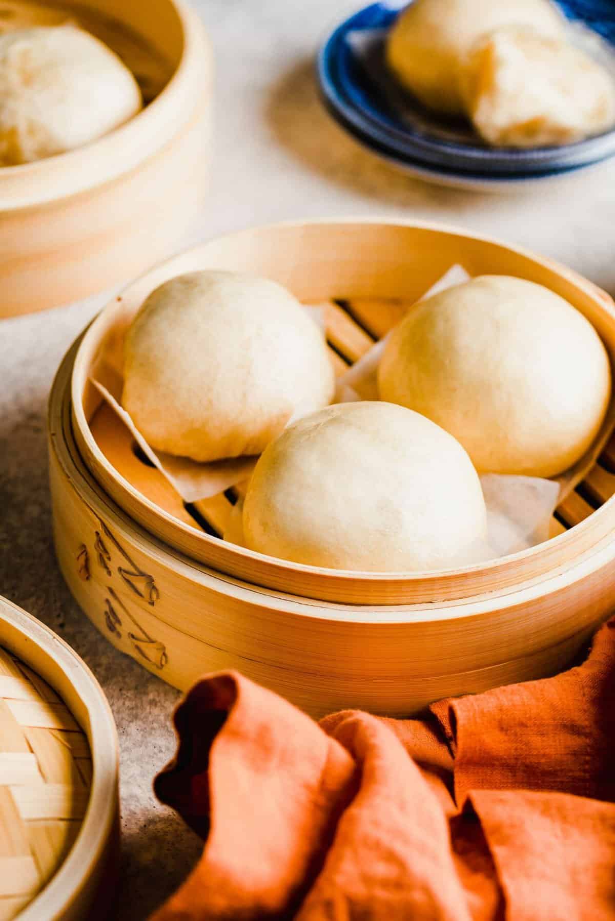 A steamer with three Chinese steamed buns in it, with another steamer of buns in the background, a plate with a steamed bun on it in the background, and a napkin in the foreground.