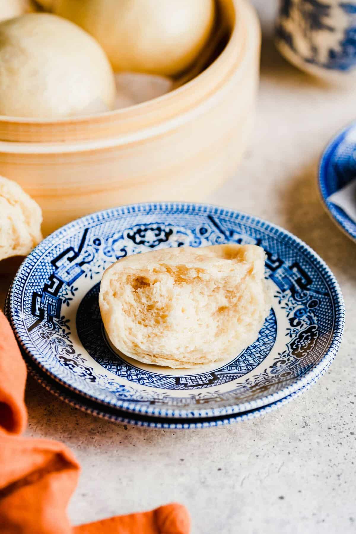 A half-eaten Chinese steamed bun on a plate, with a steamer full of buns in the background 