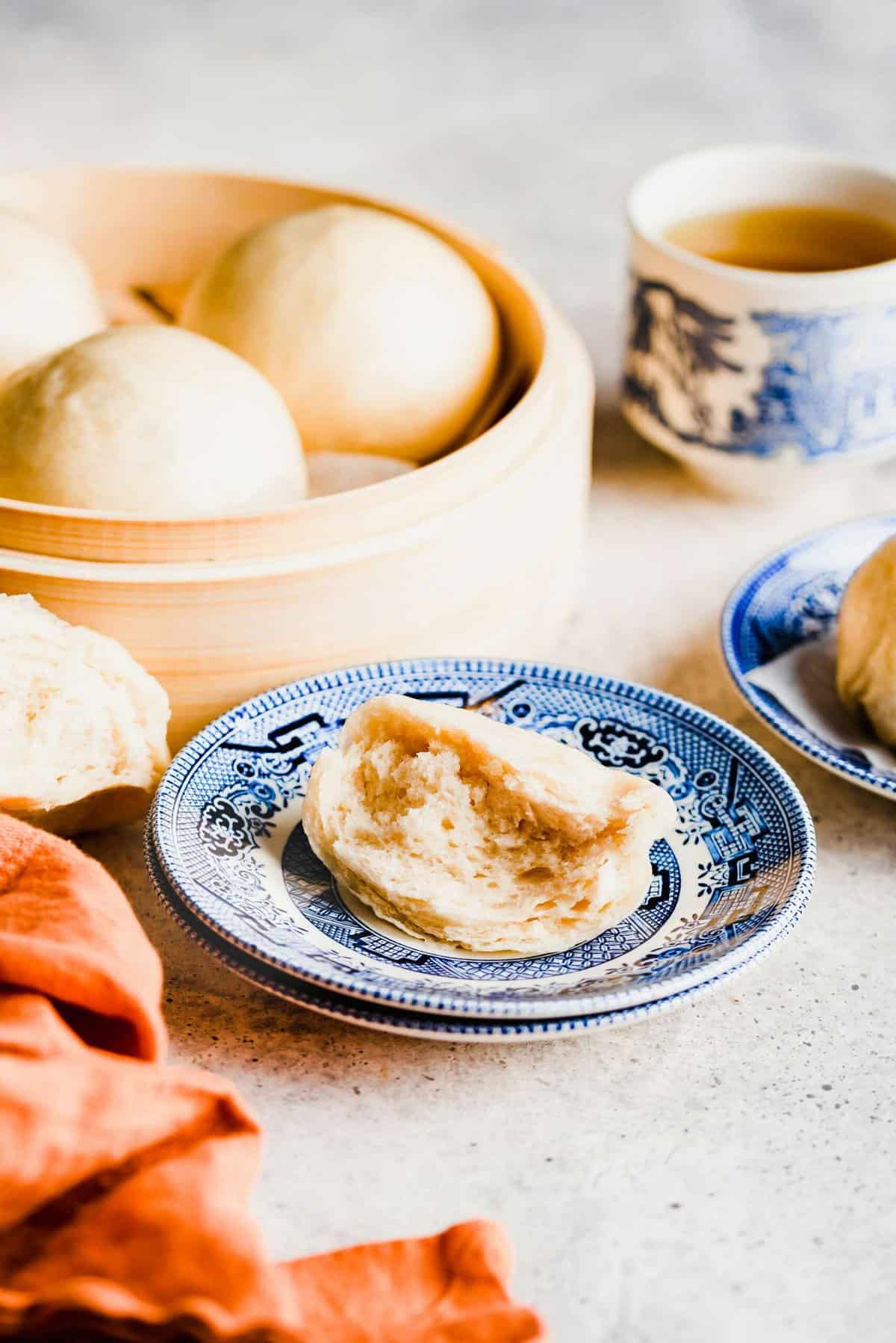 Half of a Chinese steamed bun on a plate, next to another plate, with a steamer full of buns and a cup of tea in the background