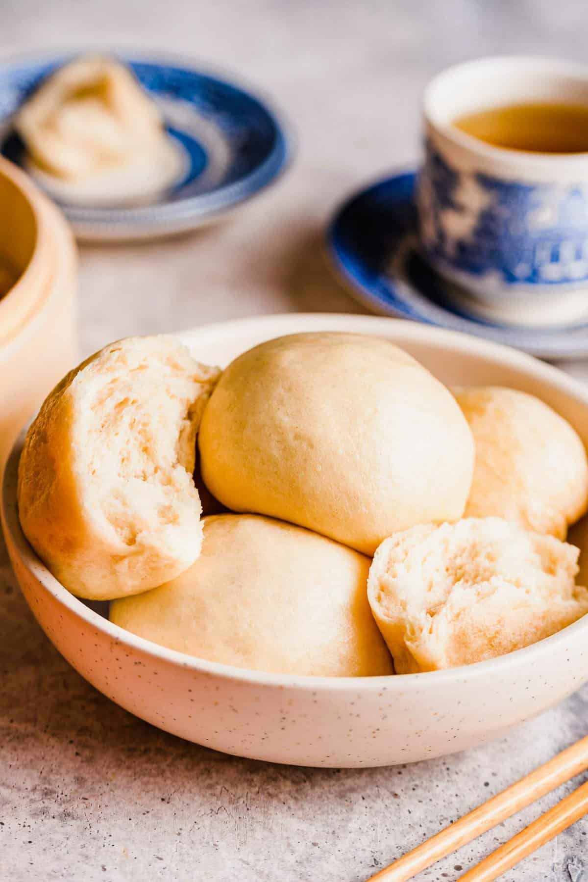 A bowl full of Chinese steamed buns, including one that's torn in half, with a cup of tea in the background