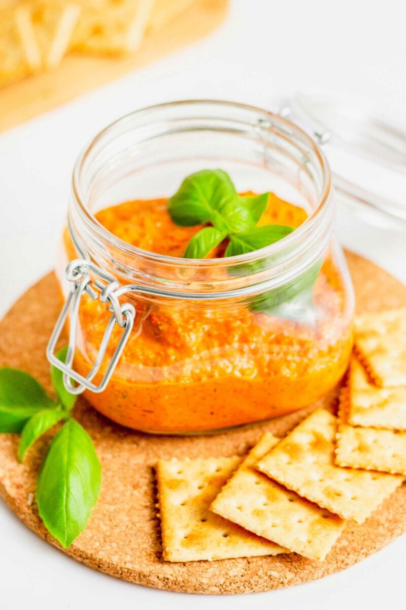 roasted red pepper pesto topped with basil in a glass jar with saltine crackers on the side