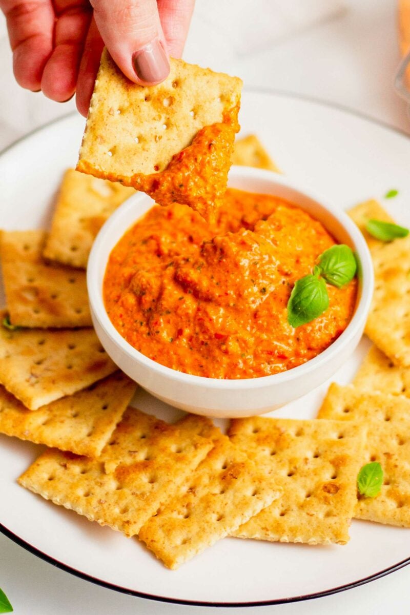 saltine cracker being dipped into roasted red pepper pesto