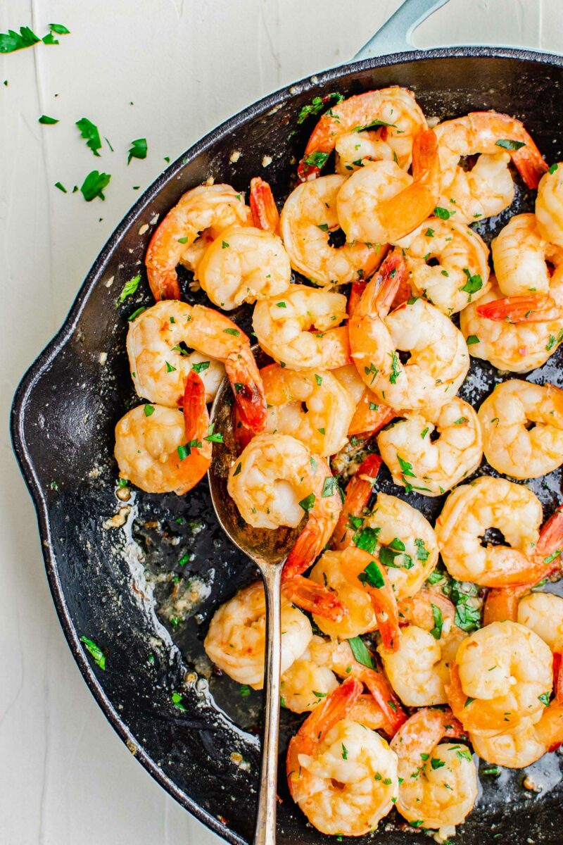 Cooked shrimp are being scooped from a black skillet with a metal spoon.