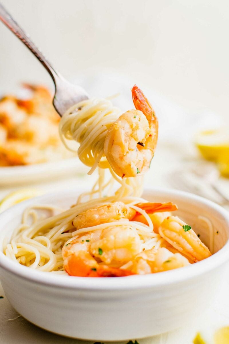 A fork is lifting a bite sized portion of noodles and a single cooked shrimp from a white bowl. 