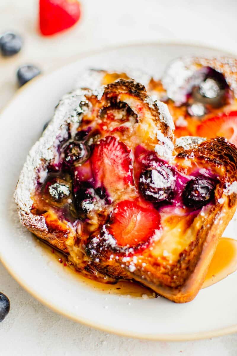 Cooked blueberries and strawberries are presented on a slice of maple syrup covered custard toast.