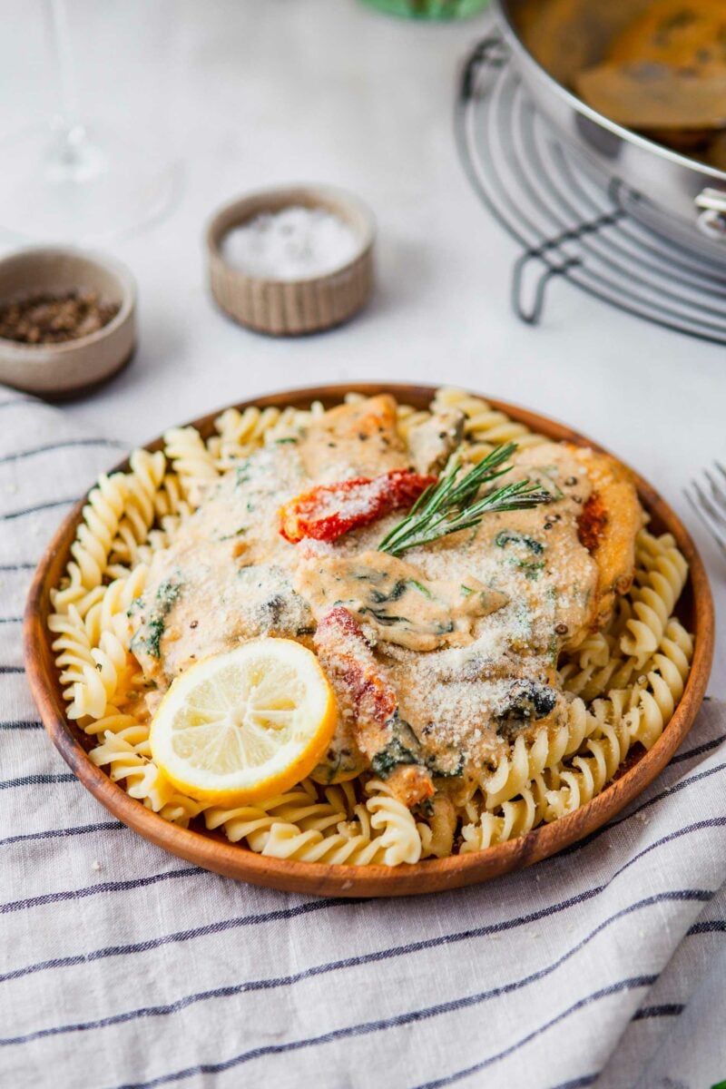 A wooden plate is topped with rotini noodles, chicken, sauce, parmesan cheese, and a slice of lemon. 