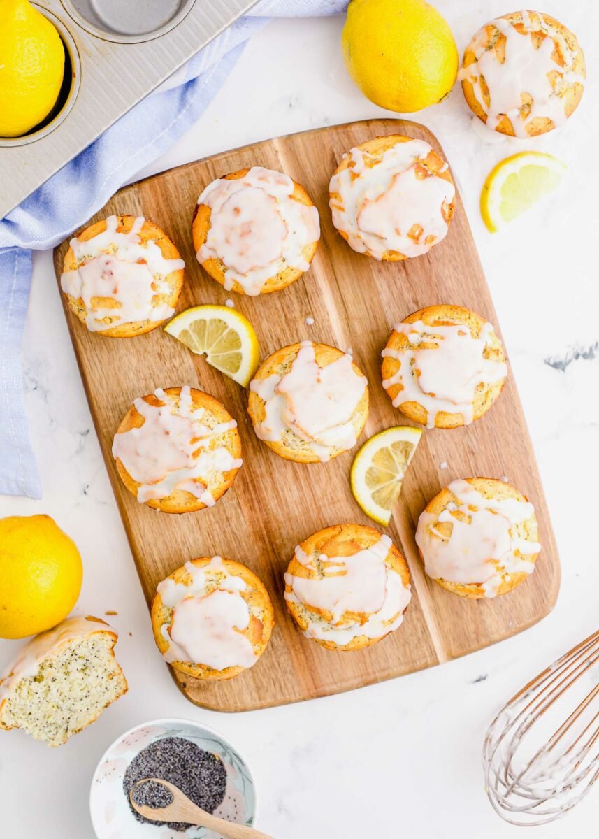 Nine glazed muffins are placed on a wooden board with two lemon slices. 