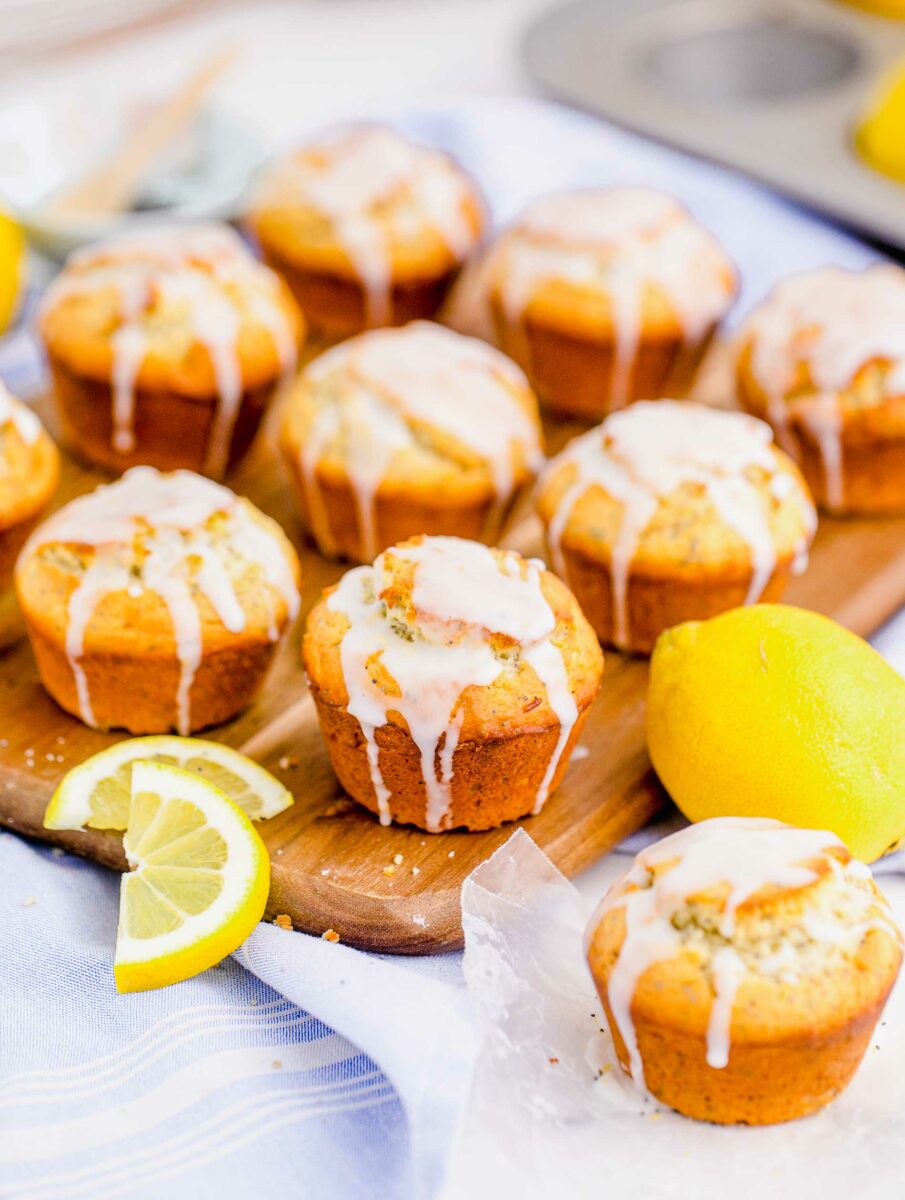 A batch of lemon poppyseed muffins are glazed and placed on a wooden serving board with whole lemons and extra muffins scattered around.