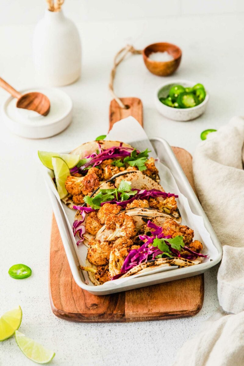 Tacos on a metal tray are placed on a wooden cutting board. 