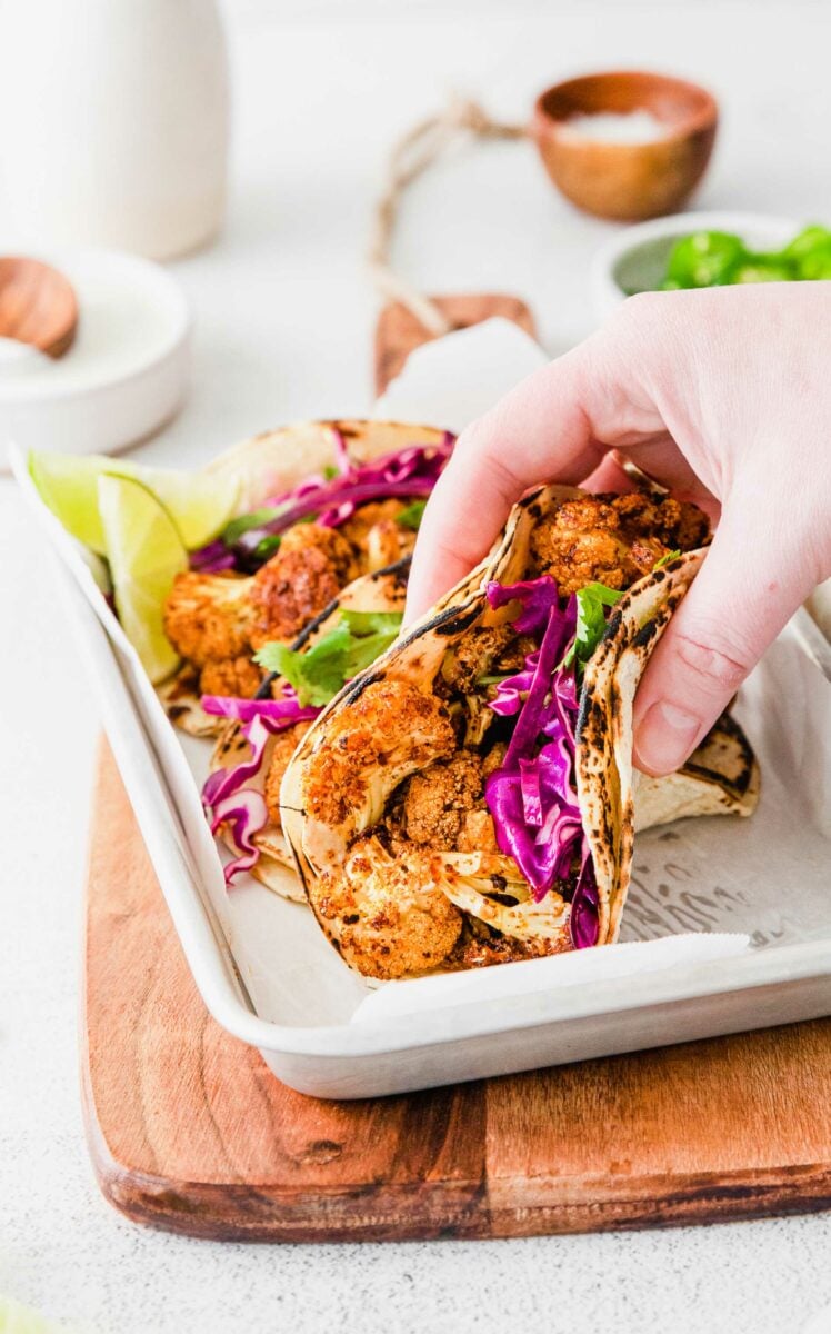 A hand is grabbing a taco from the metal platter.