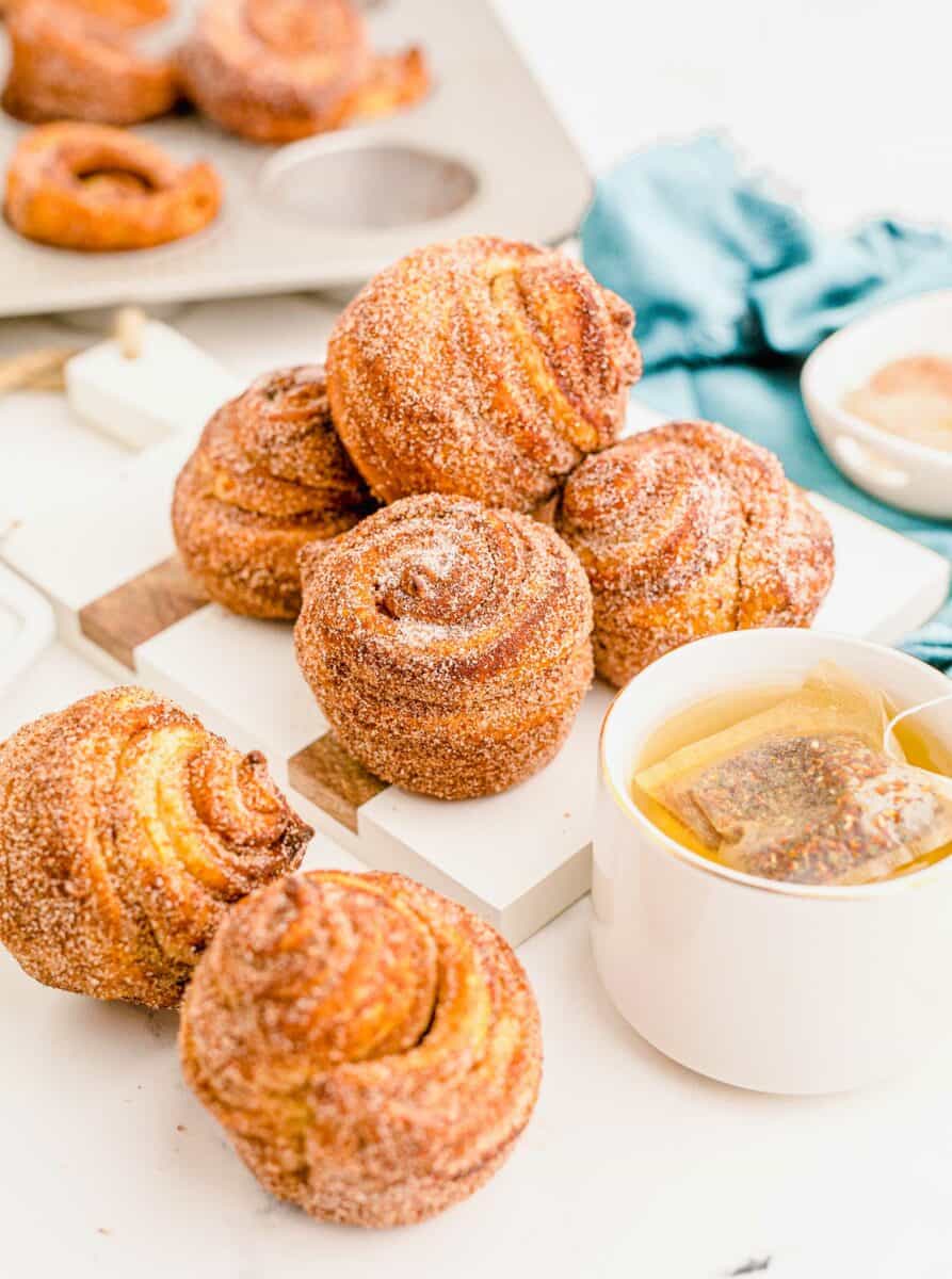 A cup of tea is placed next to a batch of cruffins on a white surface.