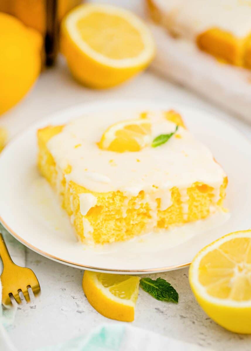 A piece of lemon sheet cake is presented on a white plate next to a lemon wedge and a fork. 