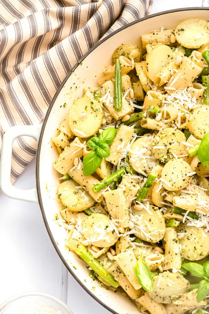 A pot filled with pesto pasta with green beans and potatoes is garnished with pine nuts, parmesan cheese, and fresh basil leaves.