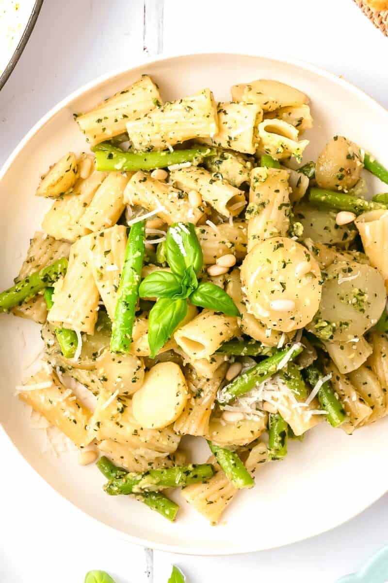 Pine nuts, parmesan, and basil are placed on top of a serving of pesto pasta on a white plate.