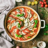 Ratatouille in round baking dish with ingredients and garnishes on tabletop