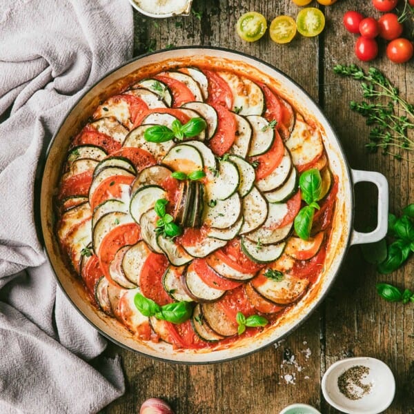 Ratatouille in round baking dish with ingredients and garnishes on tabletop