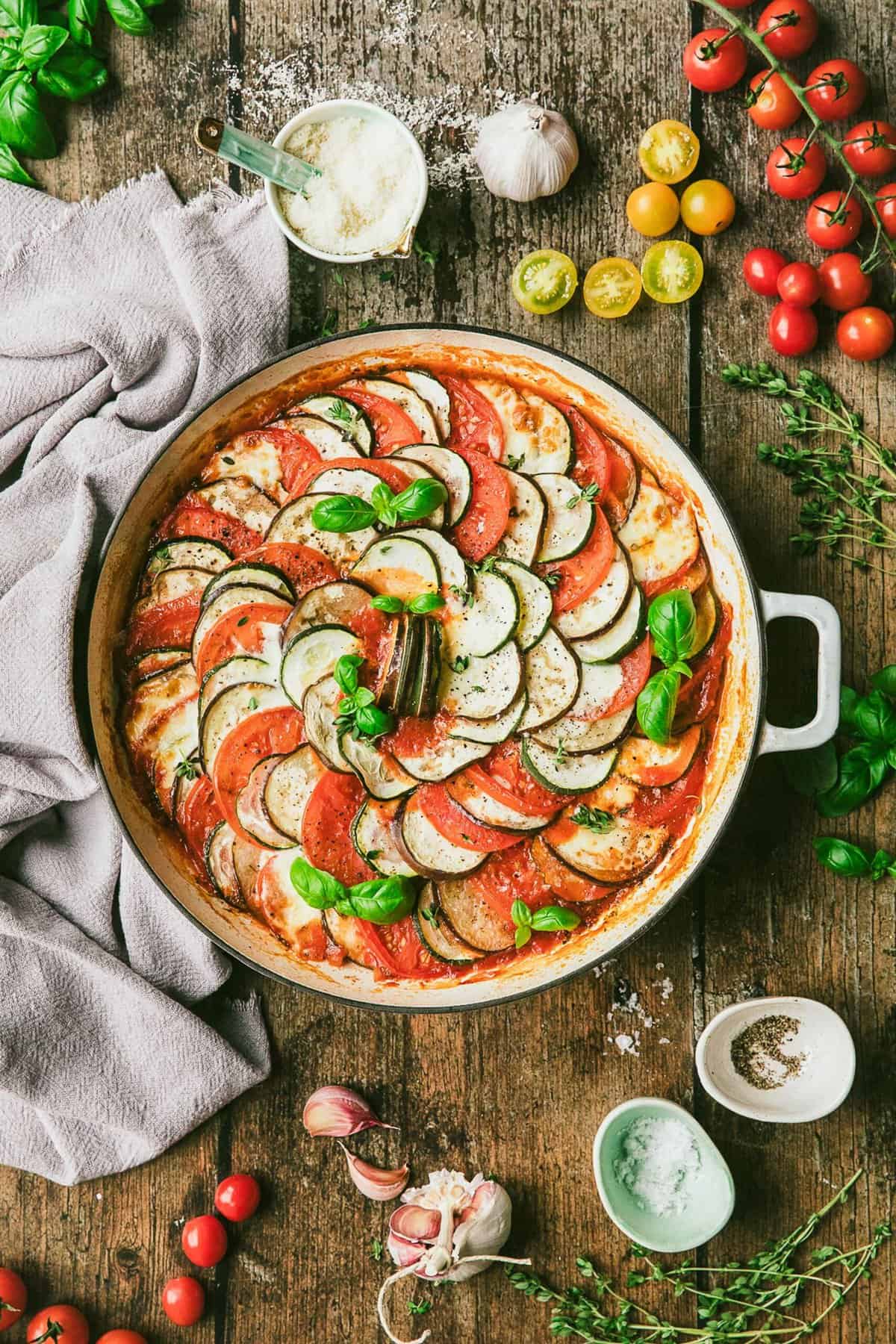Ratatouille in round baking dish with ingredients and garnishes on tabletop