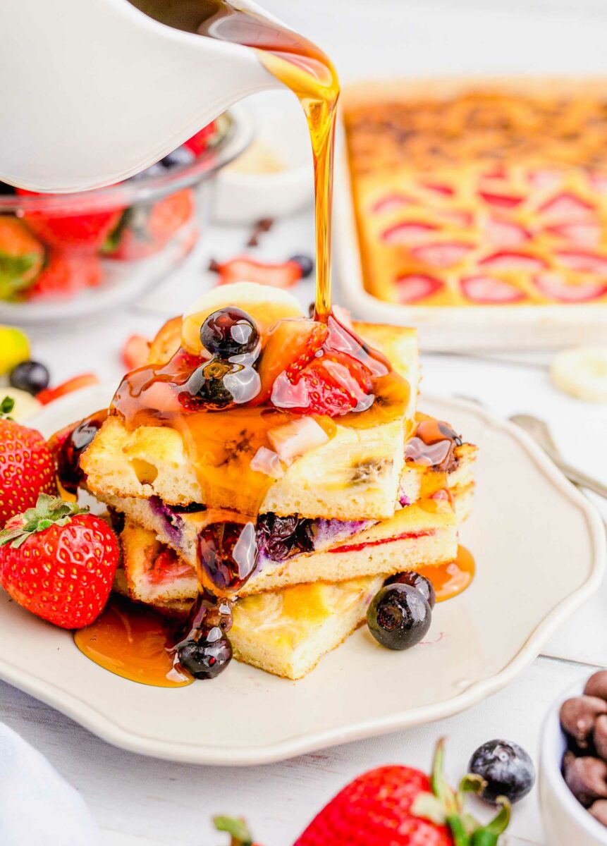 Syrup is being poured onto a stack of fruity pancakes on a white plate.
