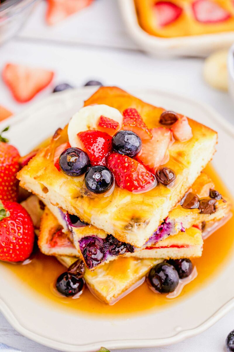 Four pancake slices are stacked on a white plate with maple syrup and extra berries. 