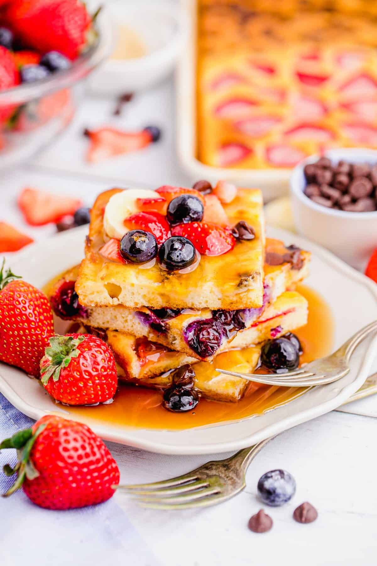 Fresh strawberries and blueberries are placed on a stack of square cut pancakes topped with maple syrup.