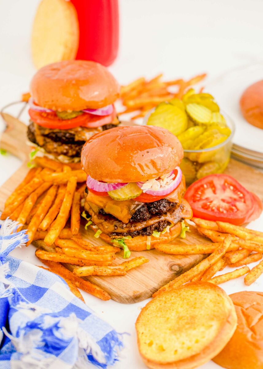 Pickles, tomatoes, and fries are placed next to two smash burgers. 