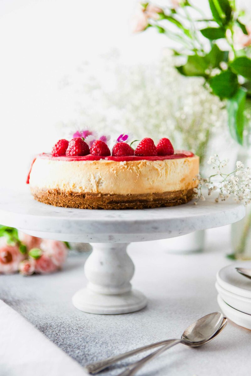 A raspberry topped cheesecake is presented on a marble cake stand. 