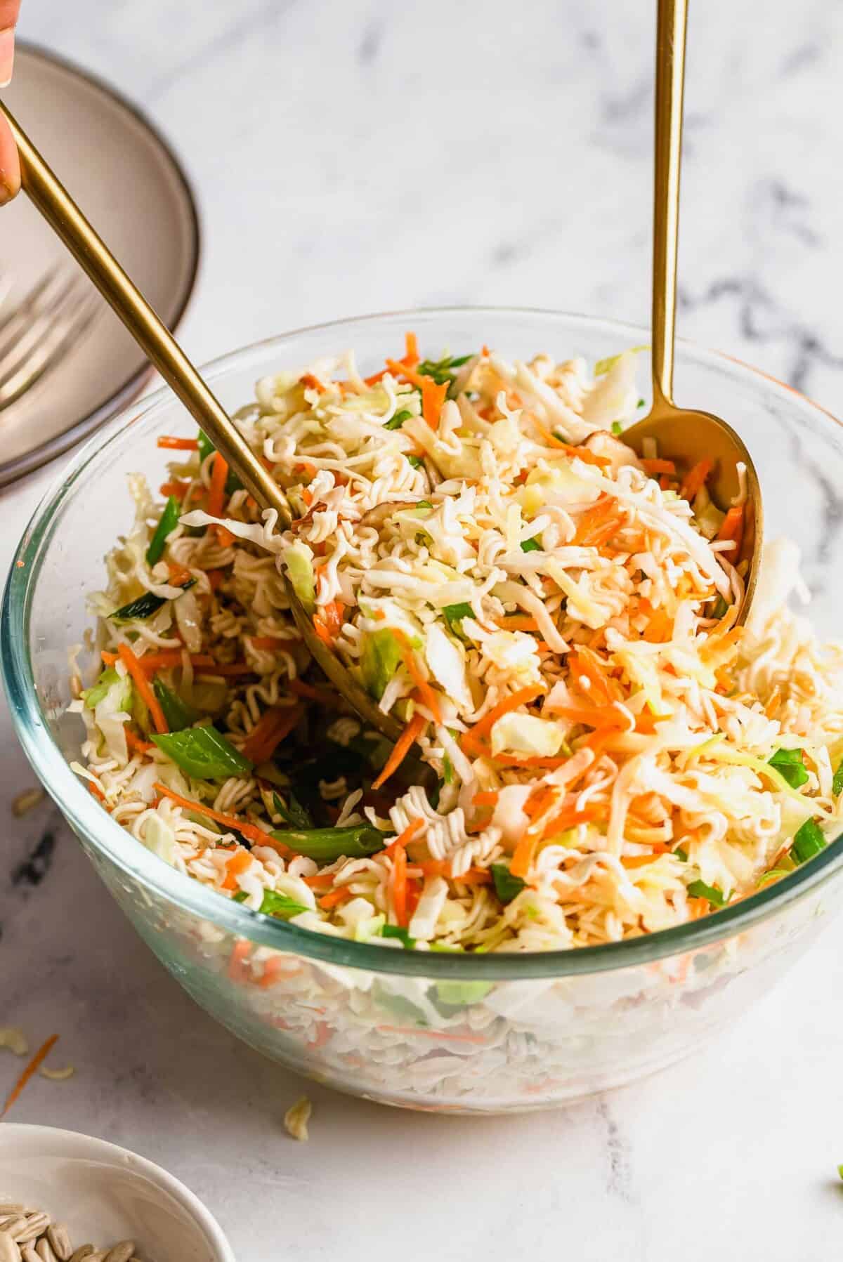 A clear bowl of Asian-inspired ramen noodle salad featuring shredded cabbage, carrots, green onions, and crunchy dry ramen noodles being mixed with gold utensils on a marble surface—perfect for your next potluck.