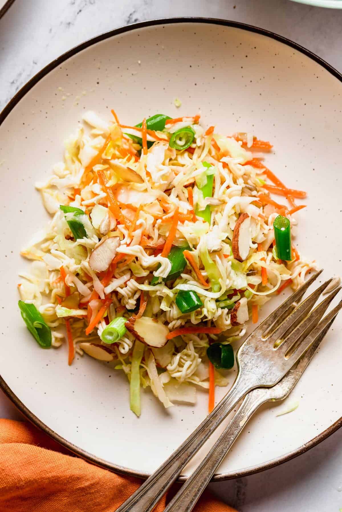 Plate of Asian ramen salad featuring crisp cabbage, shredded carrots, sliced green onions, almond slivers, and crunchy ramen noodles—all ready to serve at your next potluck, with two forks placed on the side.