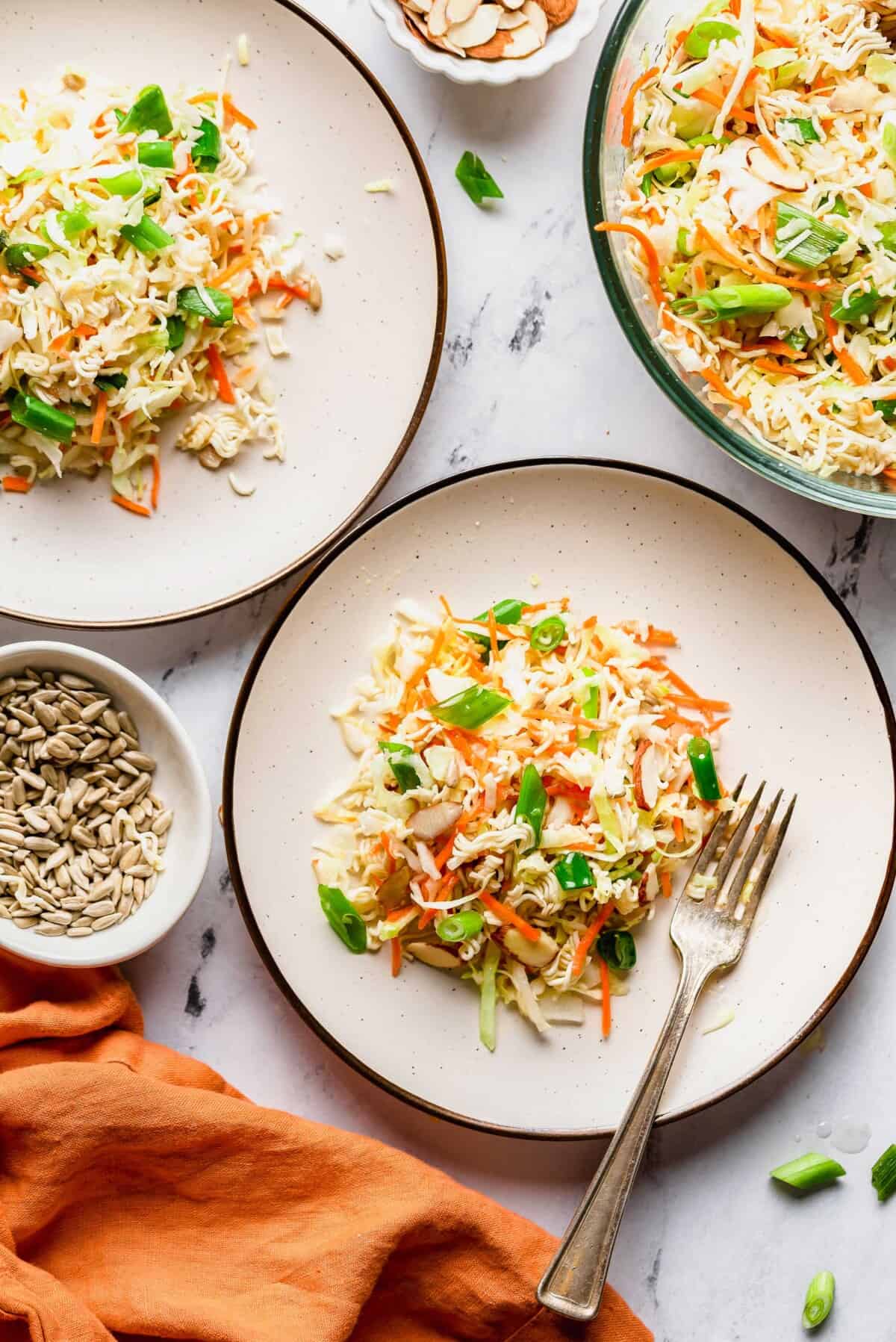 Two plates of coleslaw with shredded cabbage, carrots, green onions, and sliced almonds, next to a bowl of sunflower seeds and a serving bowl of coleslaw on a marble surface—perfect for your next potluck. For an extra twist reminiscent of asian ramen salad, try tossing in some crunchy ramen noodles for added texture and flavor.