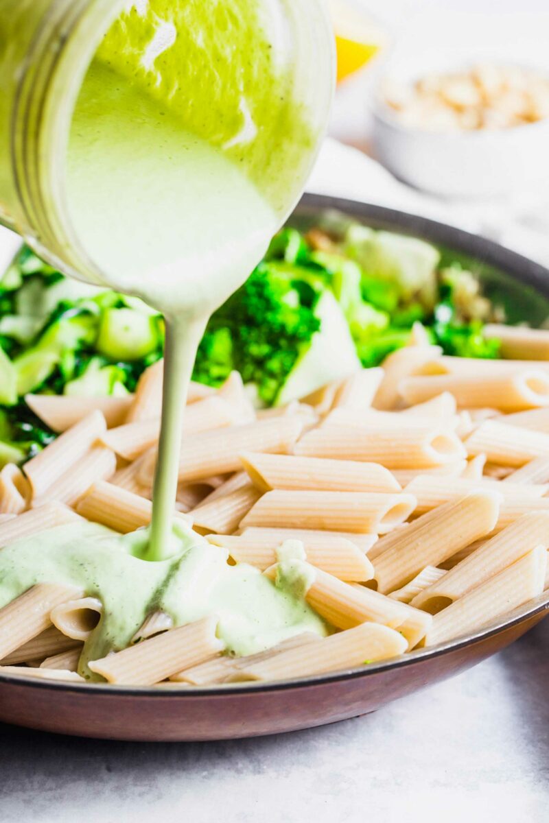 A green cashew basil pasta sauce is being poured over cooked pasta. 