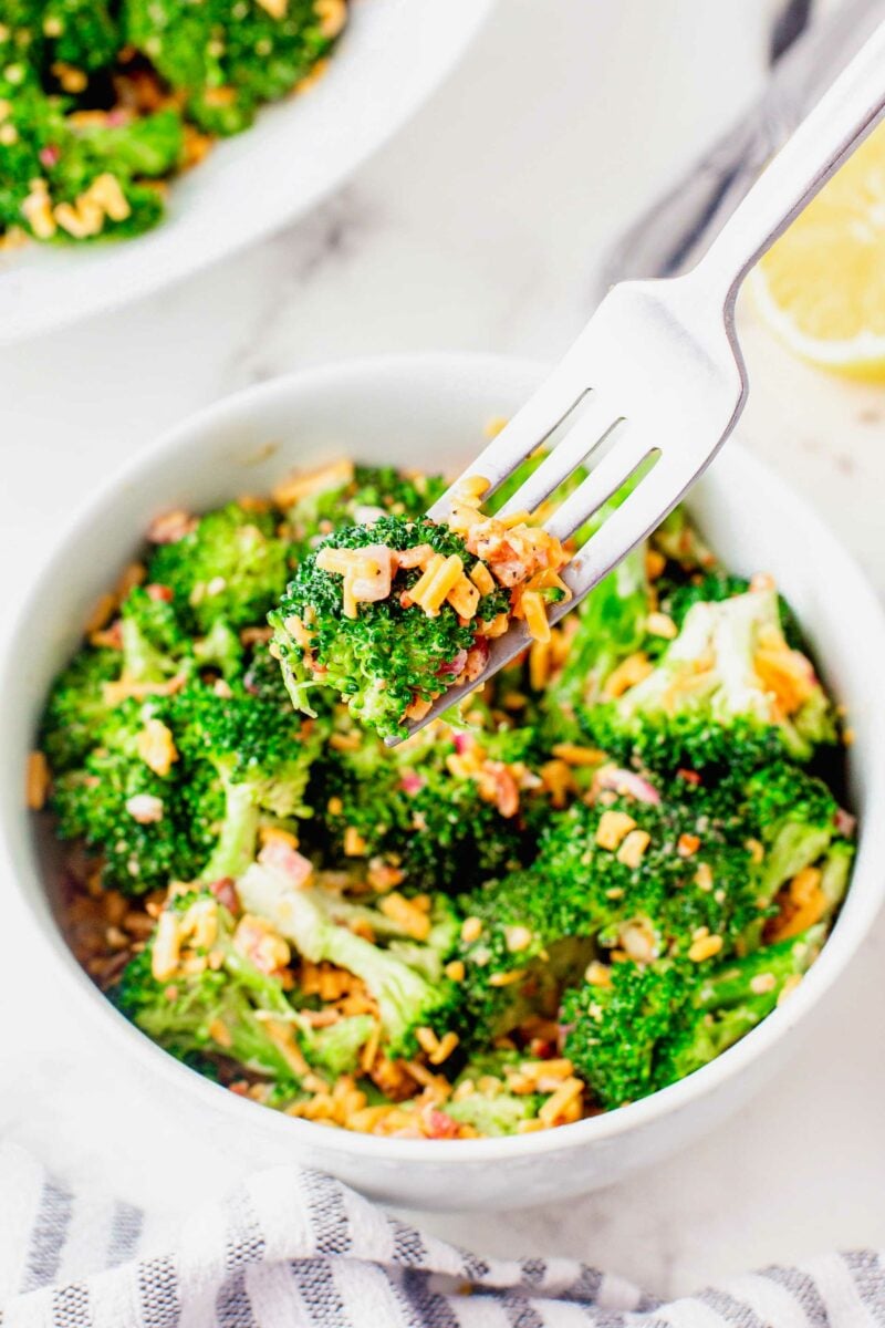 A fork has pierced a piece of broccoli above a bowl of broccoli salad. 