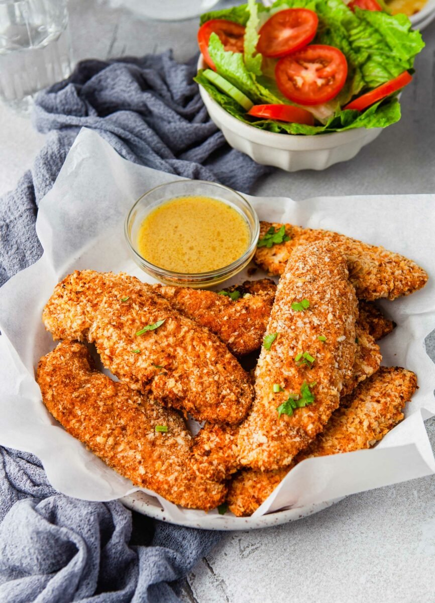 A plate lined with parchment paper is topped with baked chicken tenders and yellow dipping sauce. 
