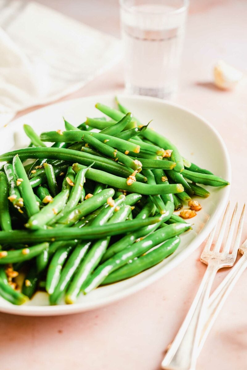 A fork and knife are placed next to a large serving of garlic and soy green beans on a white plate.