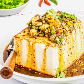 A block of tofu is topped with green onions, cilantro, sesame seeds, and a homemade oil.