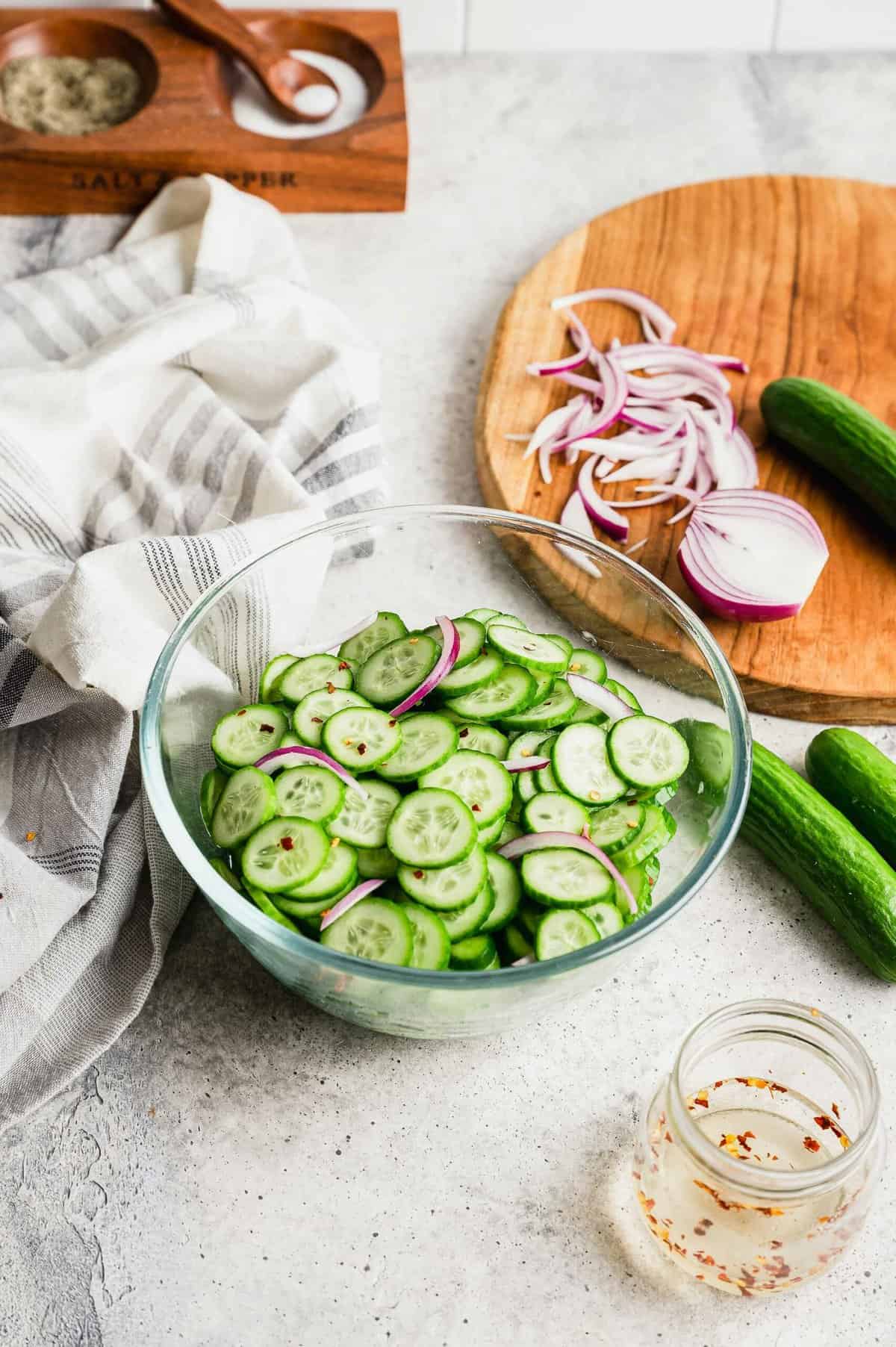 Cucumber salad in glass bowl, with cutting board in background with cucumbers and onions