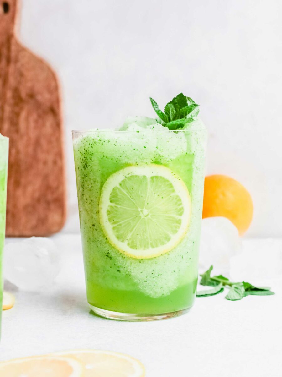 Mint leaves, ice cubes, and lemons are placed around a glass of frozen mint lemonade. 