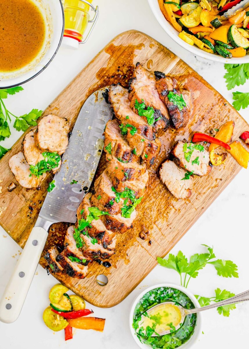 A grilled pork tenderloin has been sliced with a large knife on a wooden cutting board. 