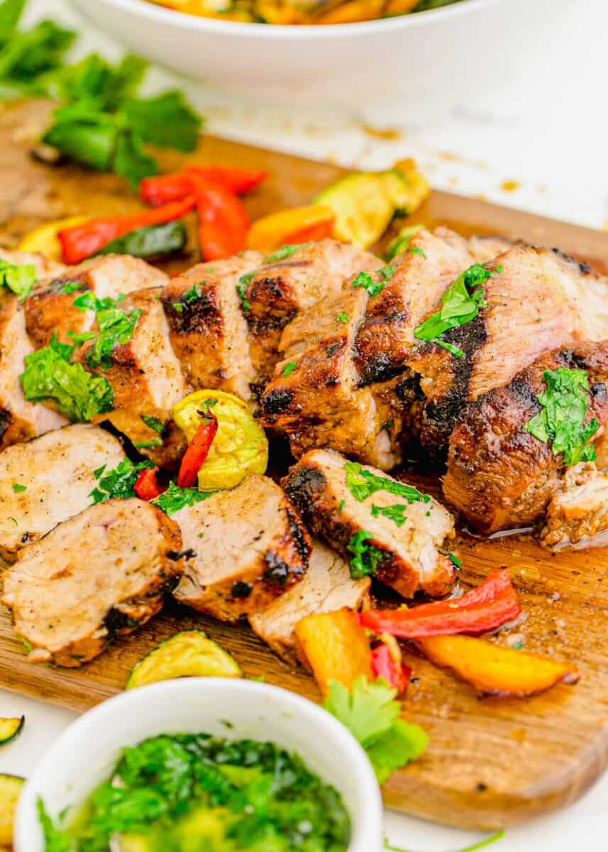 Grilled veggies are placed on a cutting board with slices of grilled pork tenderloin. 