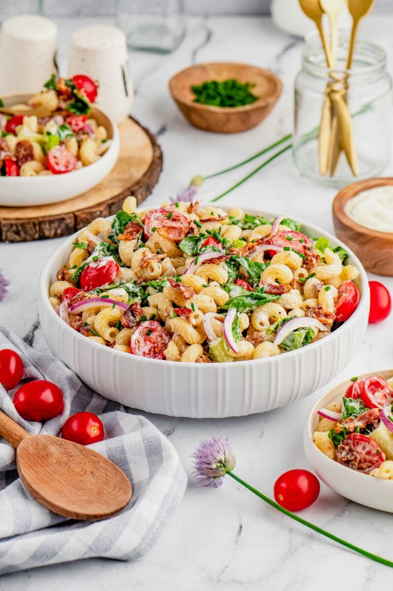 A bowl filled with BLT pasta salad is presented next to extra tomatoes, a wooden spoon, and a tea towel. 