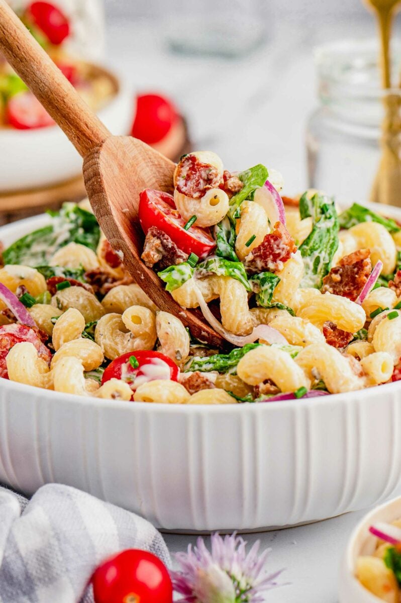 A wooden spoon is lifting a small portion of pasta salad from the bowl. 