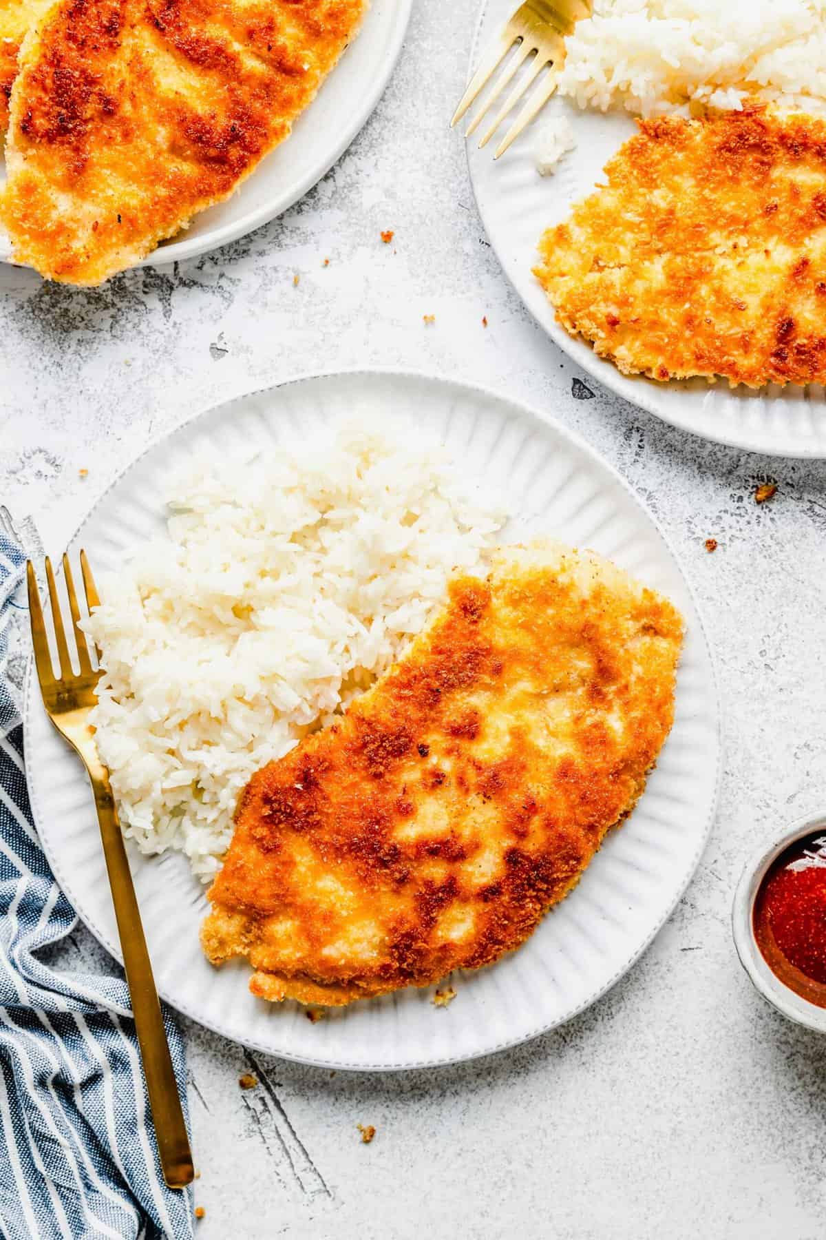 A plate with a fork, white rice, and an uncut piece of chicken katsu, next to another plate of chicken katsu and a bowl of sauce