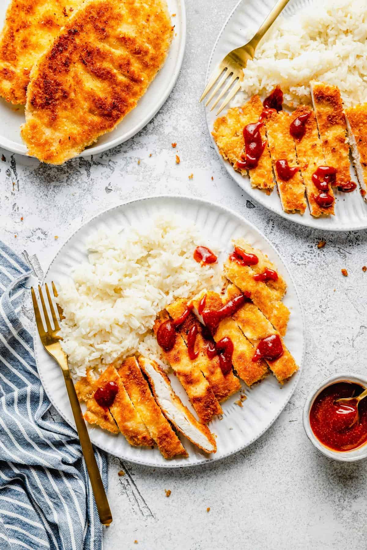 Overhead view of a plate with white rice and slices of chicken katsu topped with tonkatsu sauce, with a fork on the plate, next to another plate of chicken katsu and a serving bowl of sauce