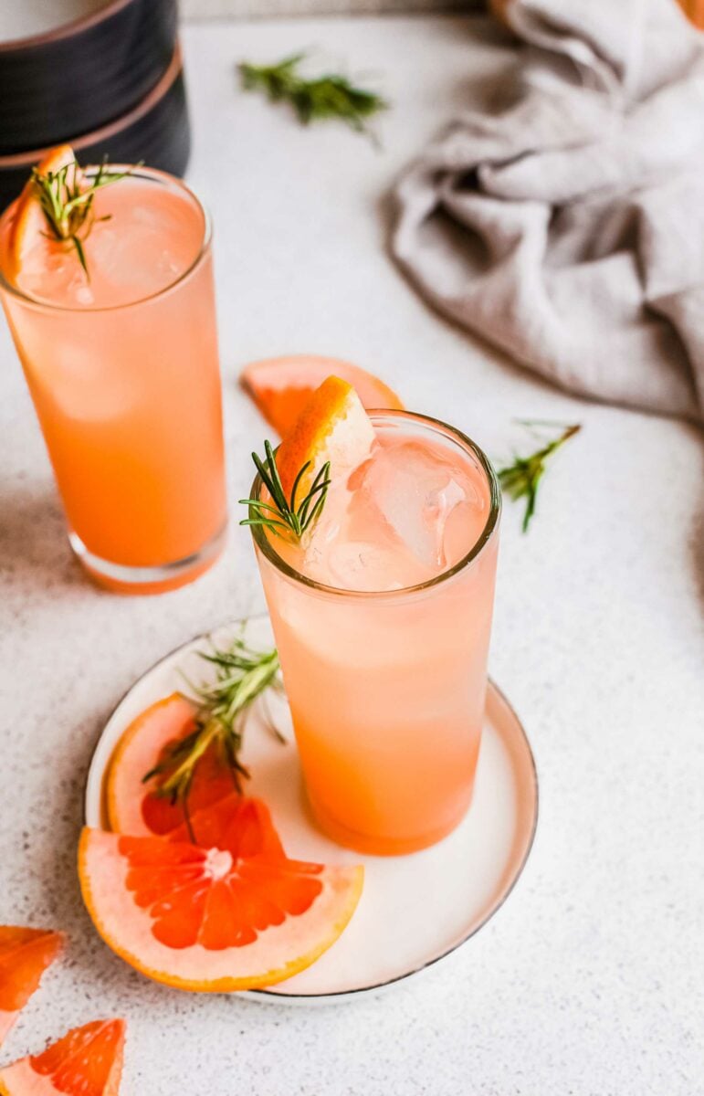 Two glasses of grapefruit gin and tonic are placed on a white surface next to extra slices of fruit. 