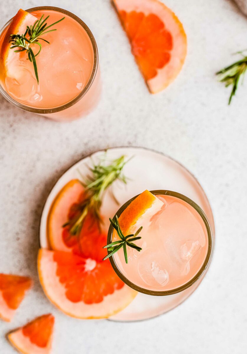 A cup of gin and tonic is placed on a white plate next to rosemary sprigs and grapefruit slices. 
