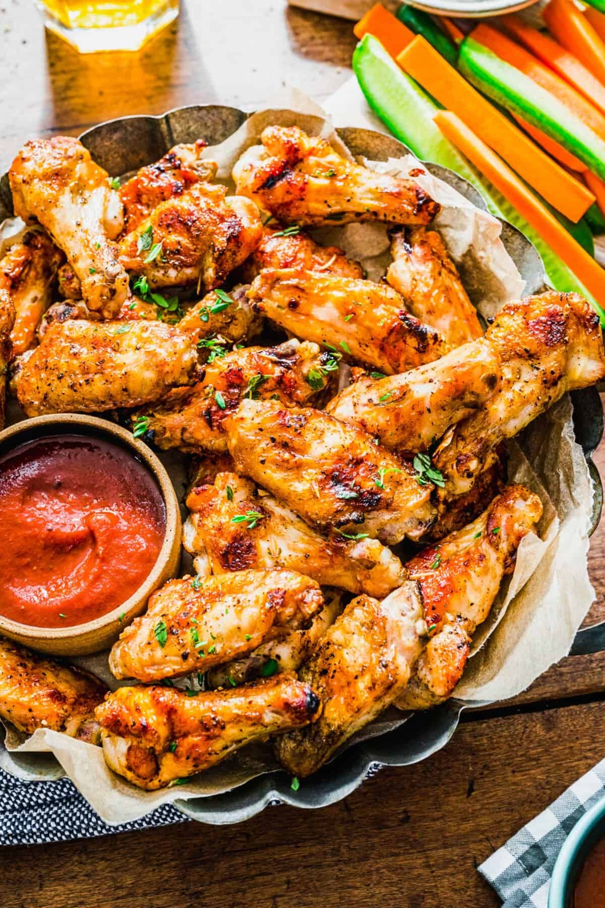 a large platter of grilled chicken wings next to a red sauce and sliced cucumbers and carrots