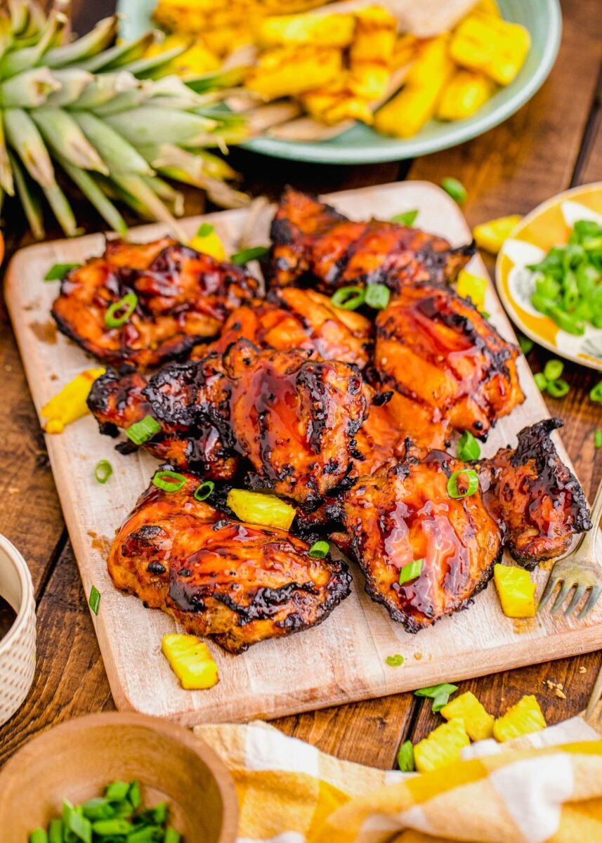 Grilled chicken thighs are placed on a wooden cutting board with garnishes. 