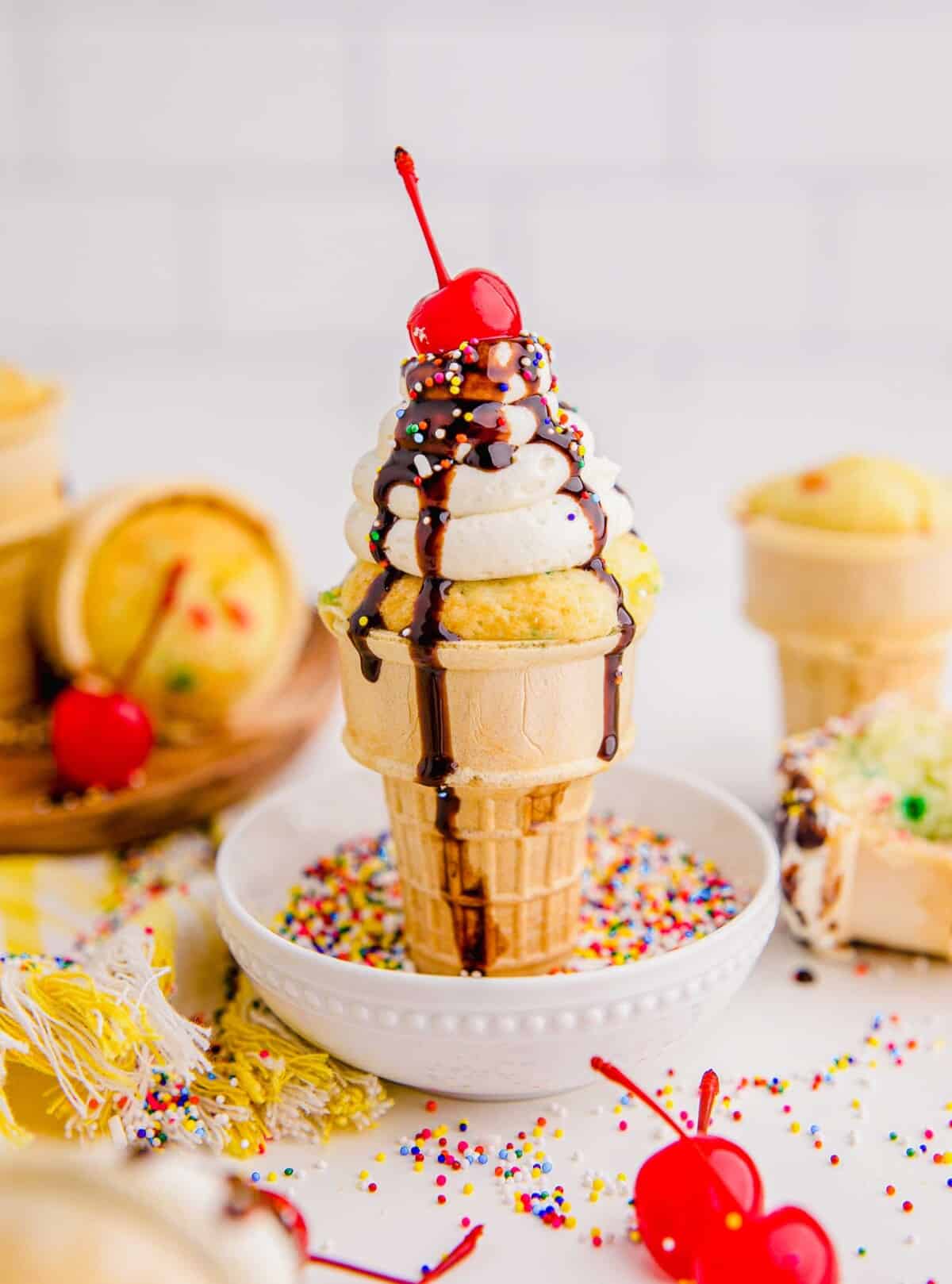 An ice cream cone cupcakes is standing in a small white bowl filled with rainbow sprinkles.