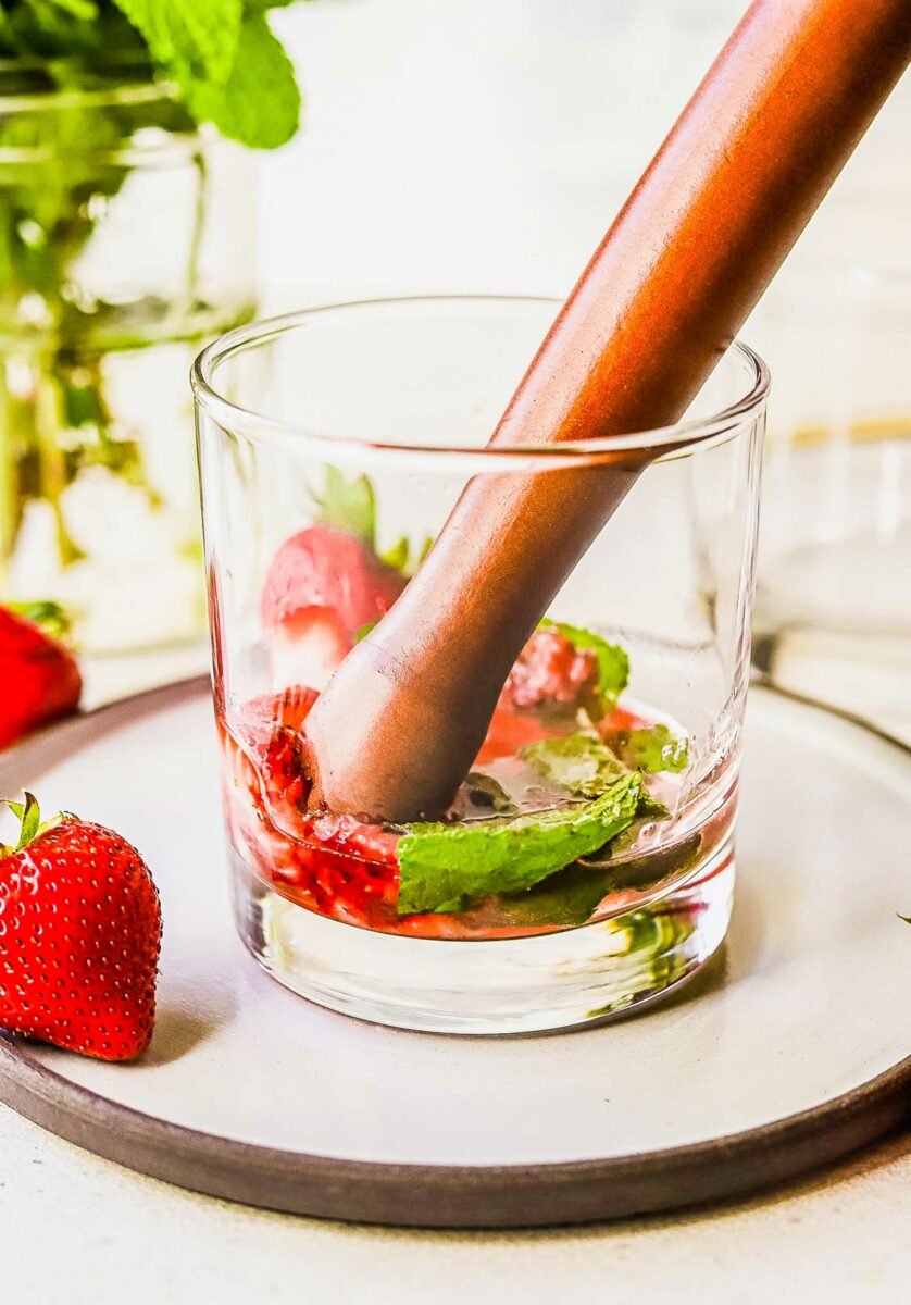 Strawberries and mint leaves are being muddled at the bottom of a glass. 