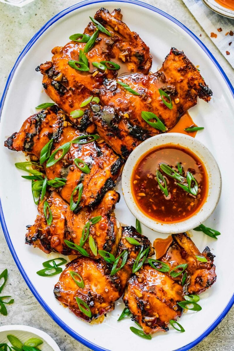 Glazed chicken thighs are placed on a white serving plate with more glaze in a small bowl. 