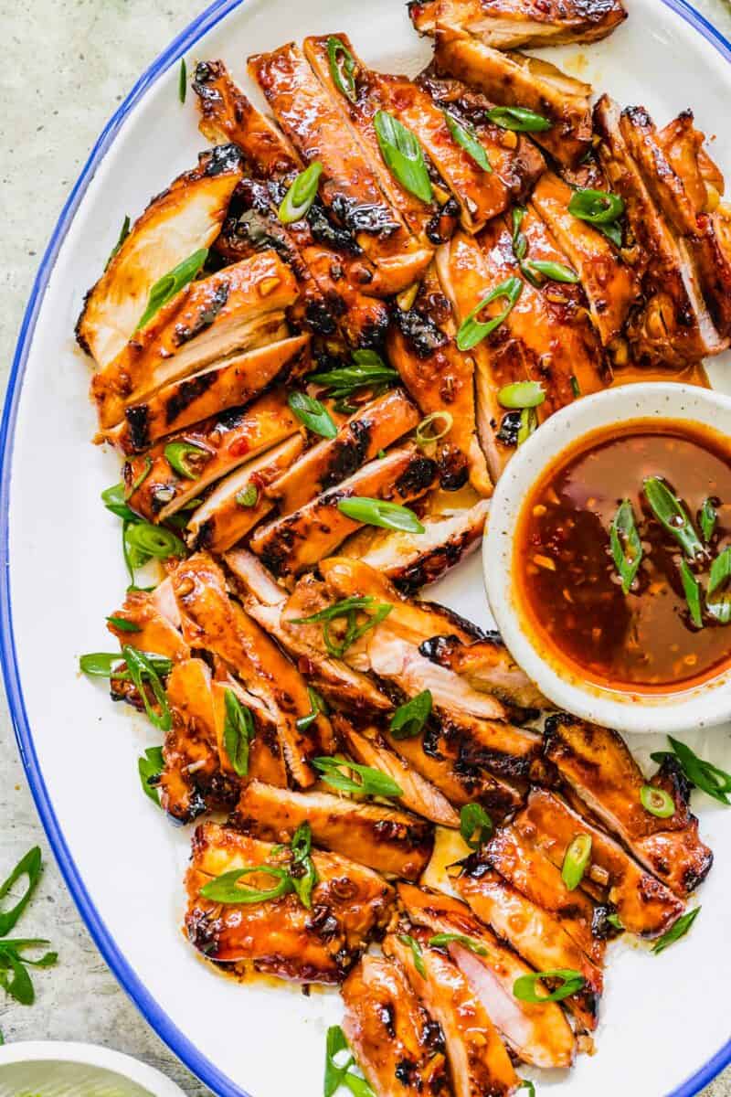 Sliced chicken thighs are served on a white plate next to a bowl of extra glaze. 